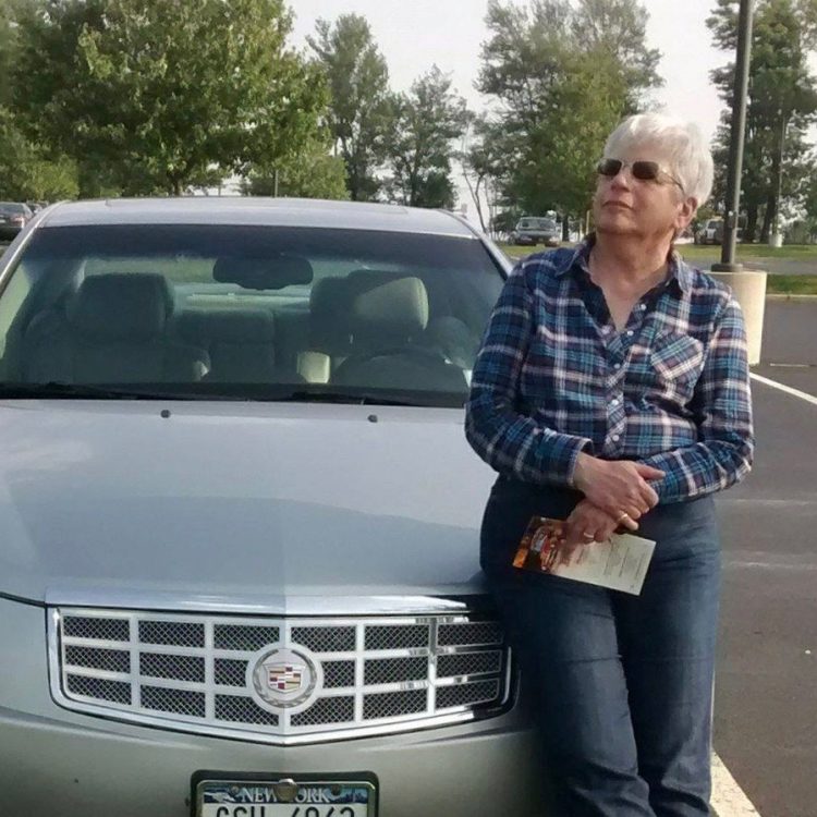 Glenna Gladstone, a white woman with short white hair, stands in front of a silver car