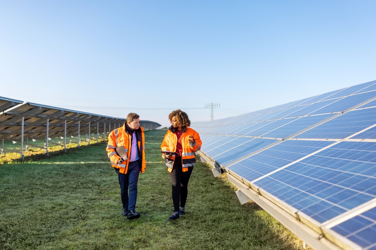 Male and female engineers working at solar power plant. Two technicians in reflective clothing walking between rows of photovoltaic panels at solar farm.