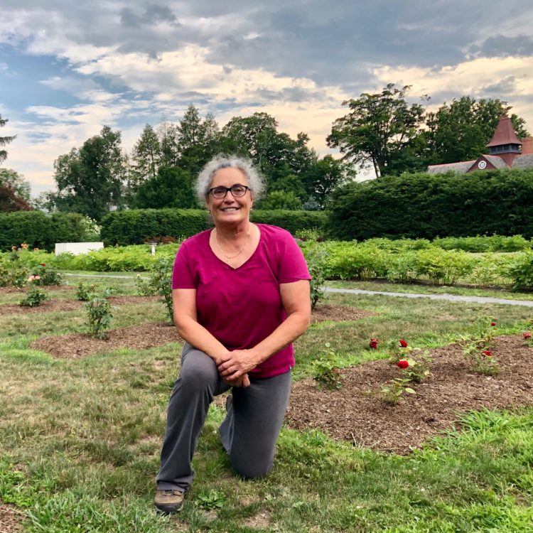 Loru Morritt, a white woman wear a bright tshirt, poses kneeling in a garden