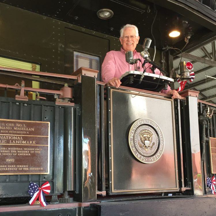 Philip Padgett, a white man wearing a red shirt, stands behind FDR's presidential podium at the FDR Library and Museum.